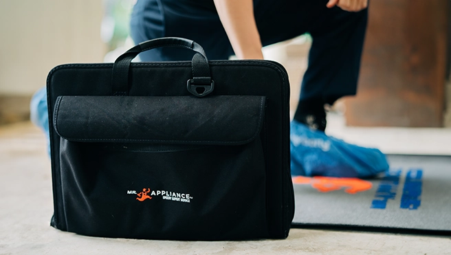 A black work bag with the Mr. Appliance logo on it and a Mr. Appliance service professional's feet, with baggies over their shoes.