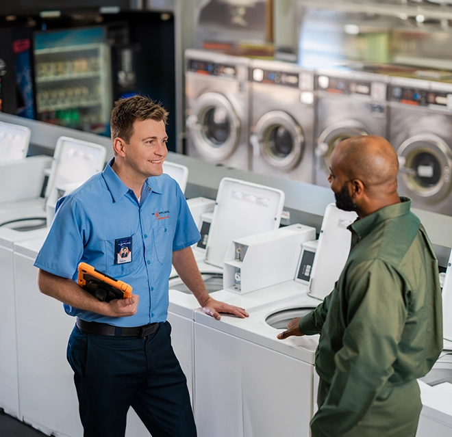 Mr. Appliance service professional smiling at customer while standing by white commercial washing machines.