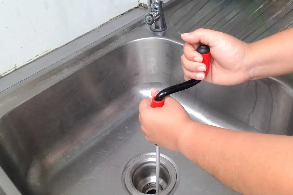 A person unclogging a kitchen sink with a tool.