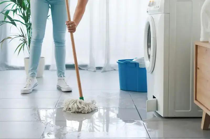 A woman uses a mop to clean up a washing machine leak on a tiled floor.