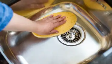 A hand wiping down a stainless steel sink with a yellow sponge.