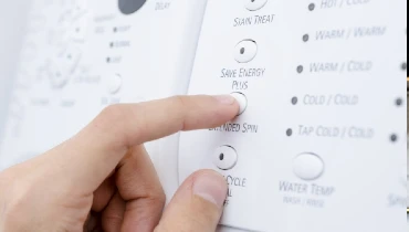 A person's finger pressing a button labeled Save Energy Plus on a washing machine.